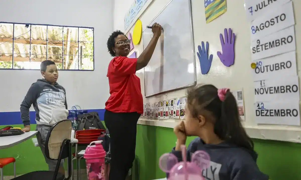 Educação com acolhimento: professora ensina em ambiente inclusivo e participativo Professora escreve no quadro branco enquanto crianças observam em sala de aula colorida.