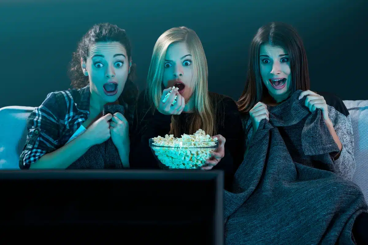 Três amigas assistindo a um filme de terror, com expressões de medo, segurando pipoca e cobertores.