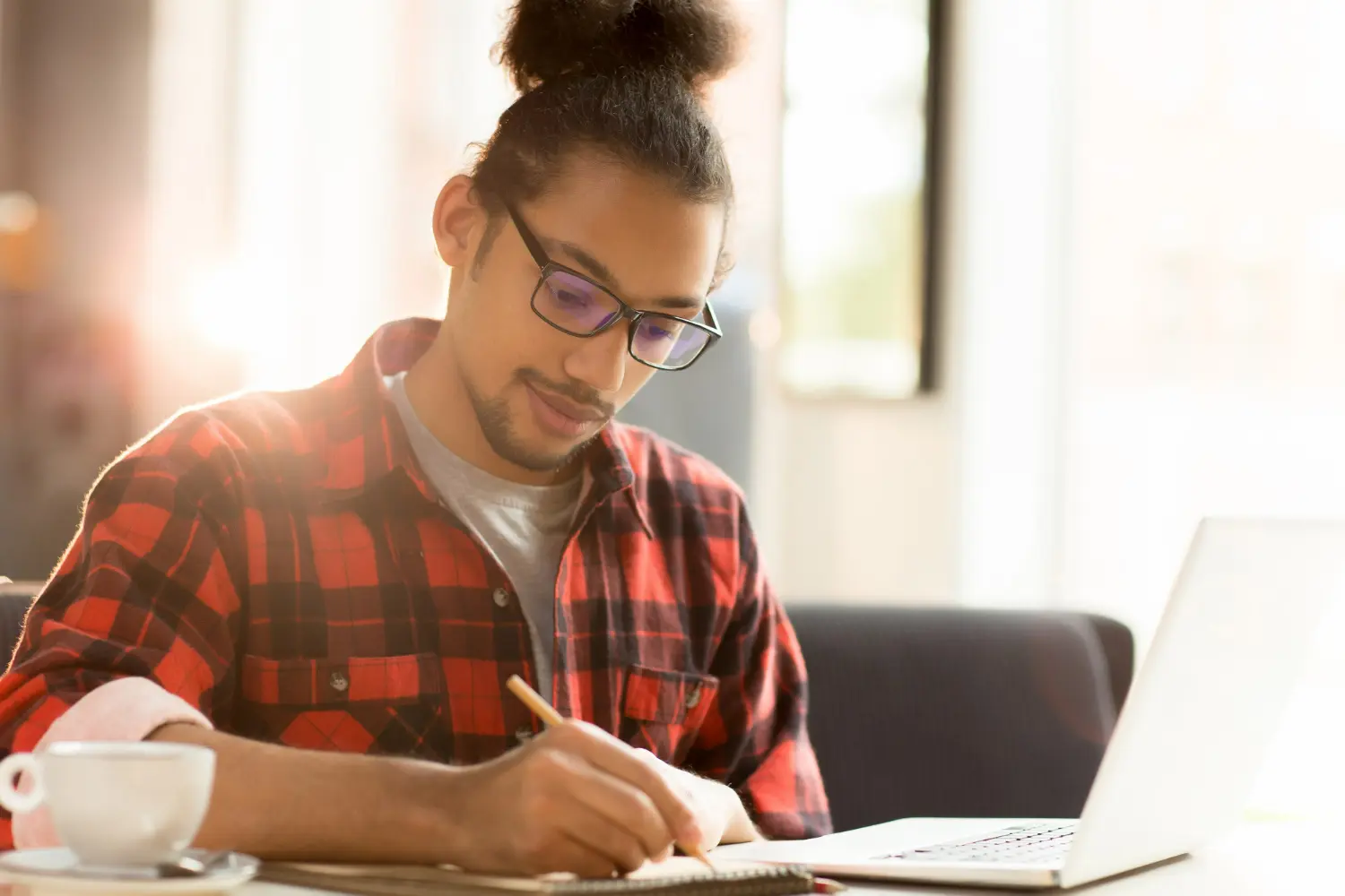 Homem escrevendo enquanto reflete sobre a etimologia de 'expressão'. Homem escrevendo em um caderno enquanto utiliza um laptop, refletindo sobre a etimologia da palavra 'expressão'.