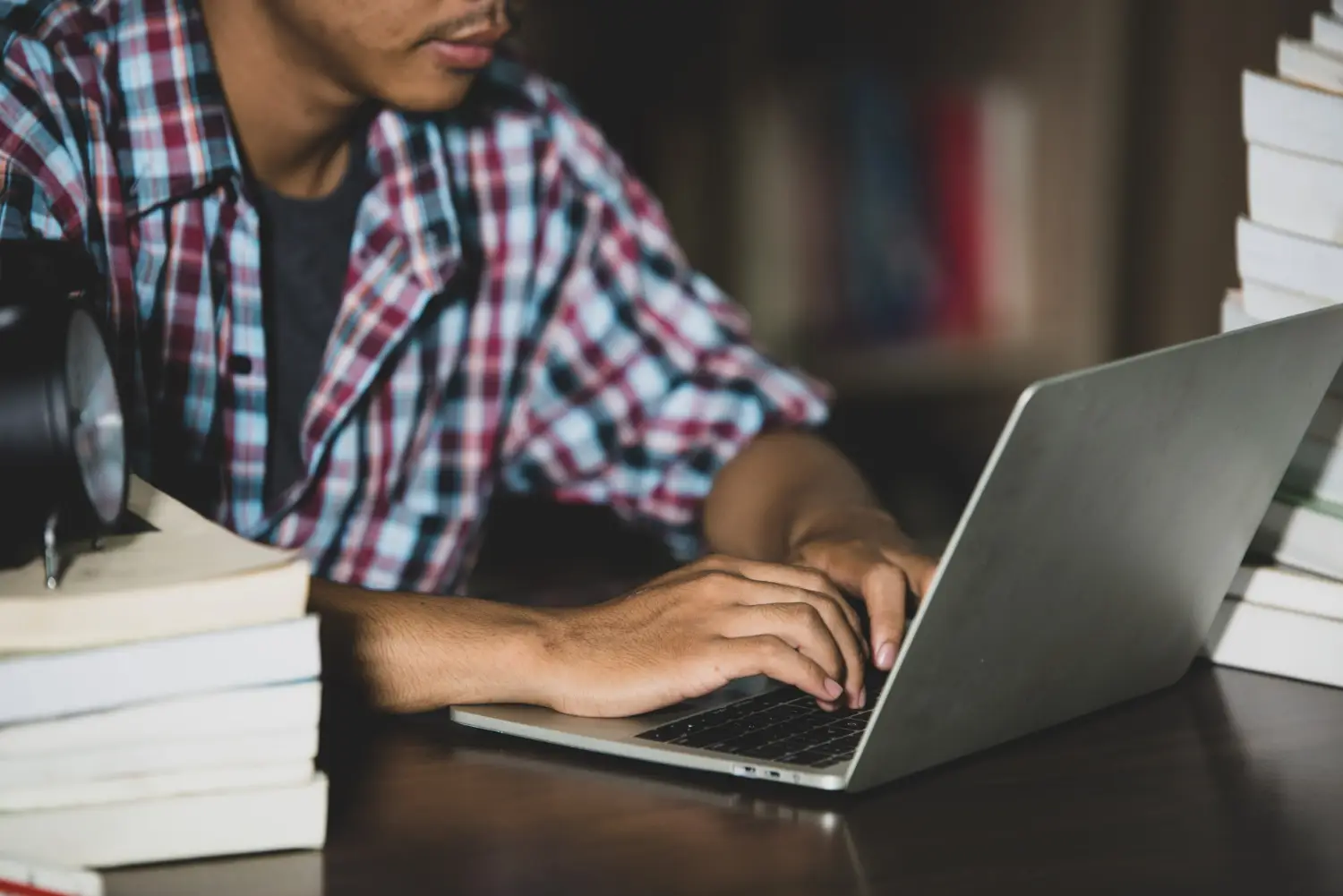 Jovem em um ambiente de estudo, digitando no laptop enquanto livros estão ao seu redor, simbolizando a dedicação aos estudos para cursos técnicos.