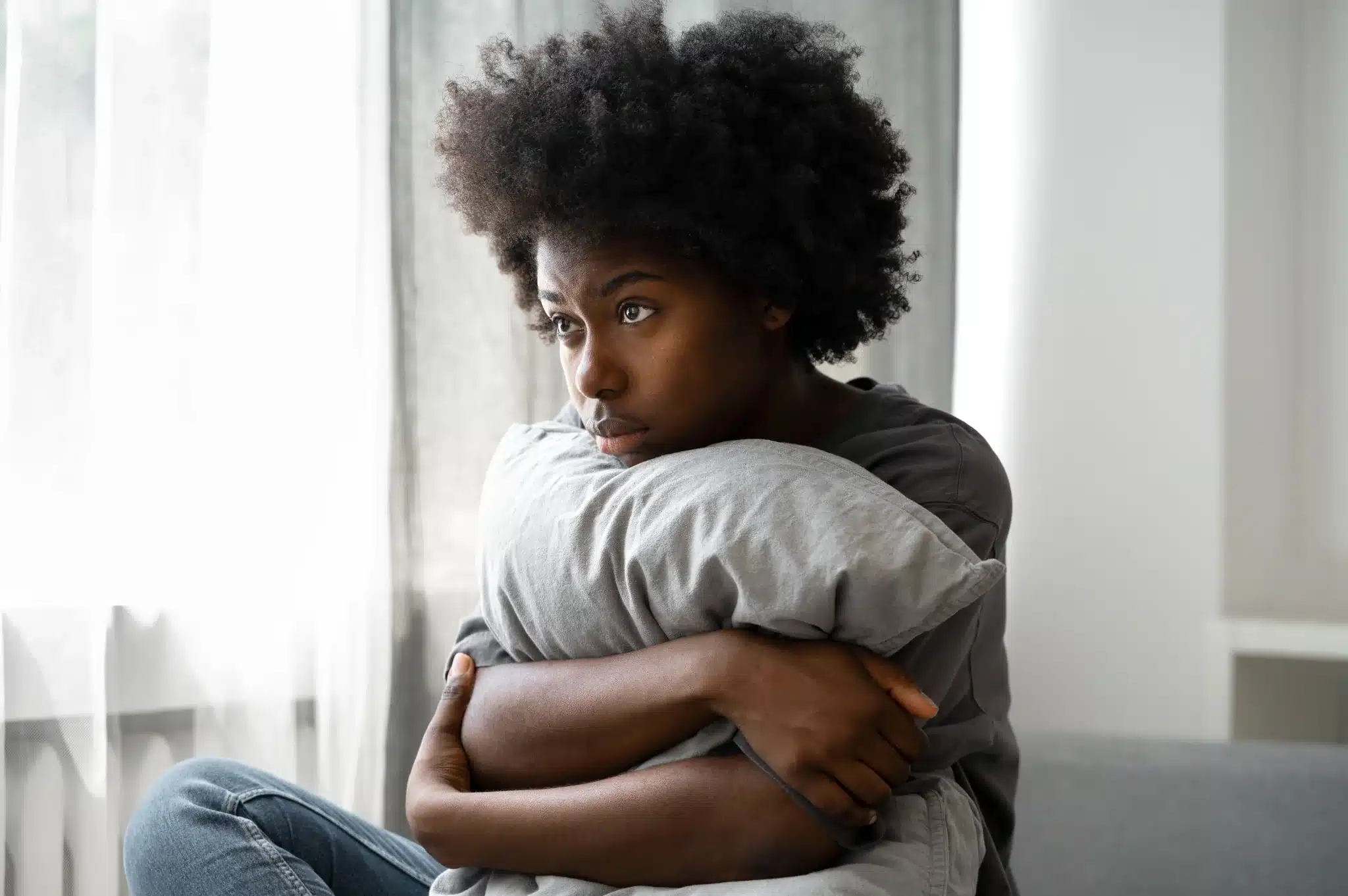 Jovem reflexivo e emocional em ambiente doméstico Jovem com cabelo afro segurando um travesseiro, com expressão pensativa e triste, sentado em um sofá em um ambiente doméstico.