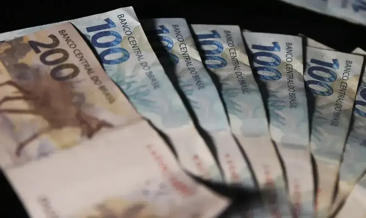 Notas de R$100 e R$200, com a logo da Caixa ao fundo.