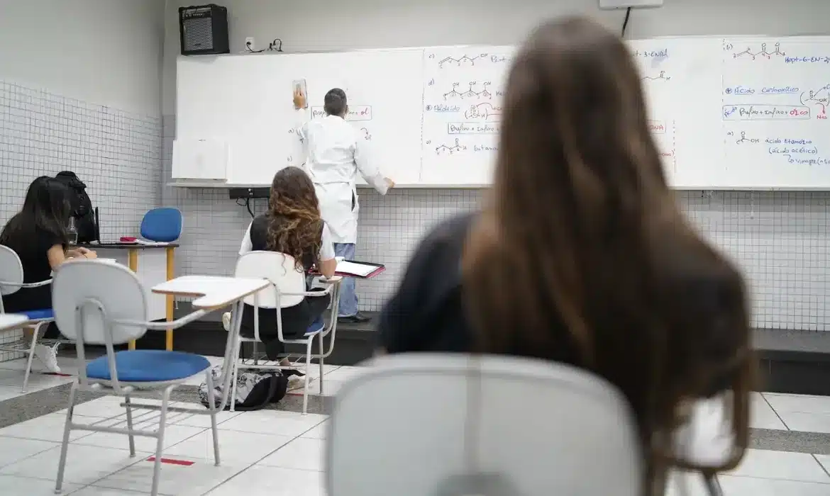 Programa Pé-de-Meia - Incentivo Financeiro para Estudantes do Ensino Médio. Alunos assistem aula em escola pública com professor escrevendo na lousa.