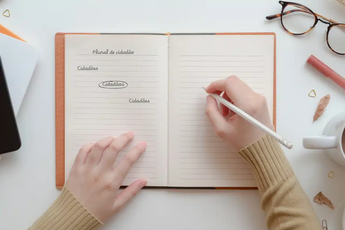 Anotação sobre o plural de cidadão em caderno aberto Mãos femininas escrevendo em um caderno aberto com anotação sobre o plural da palavra cidadão.
