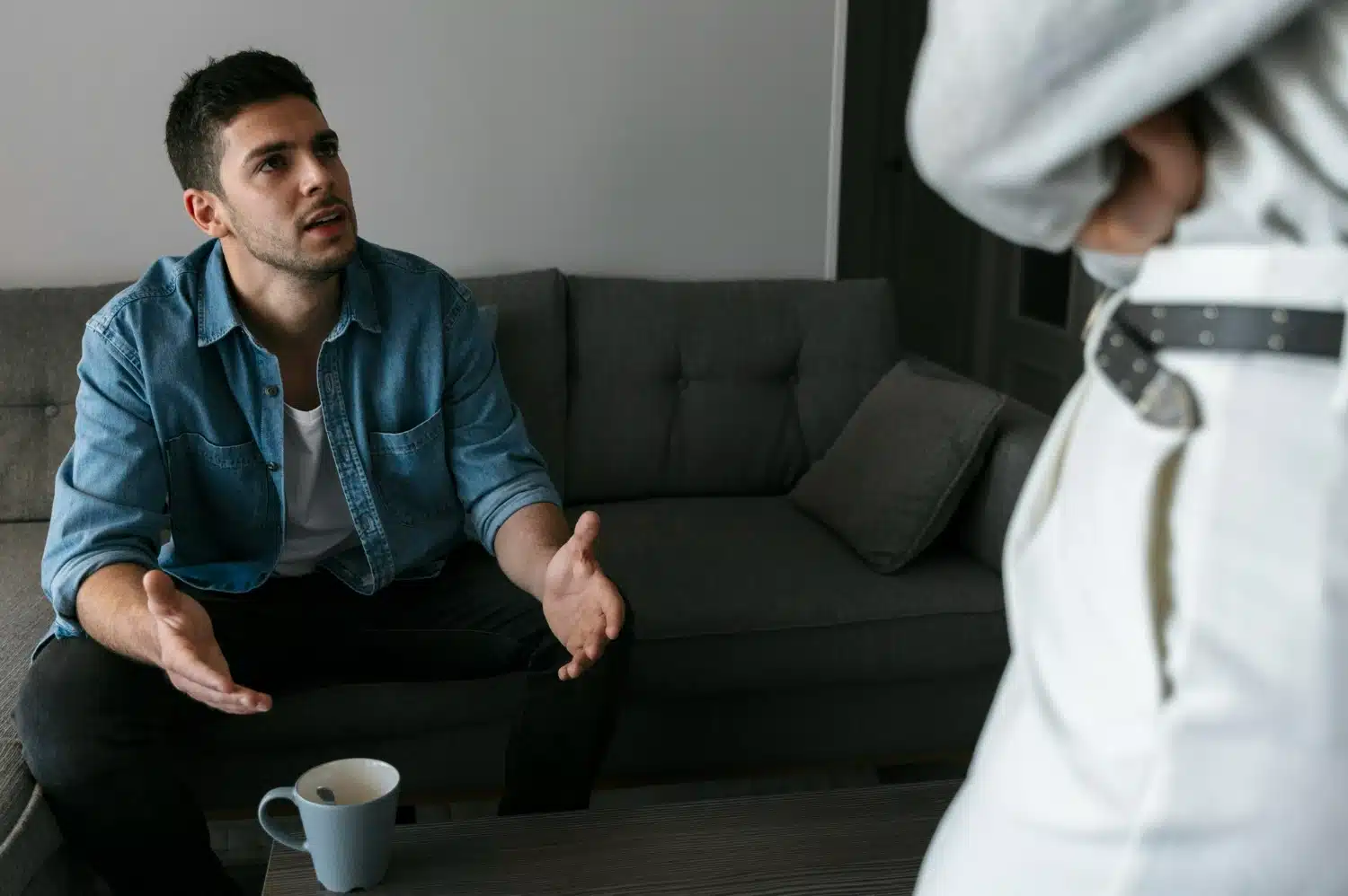 Homem sentado no sofá, conversando de forma séria com outra pessoa em pé, expressão de explicação ou argumentação.