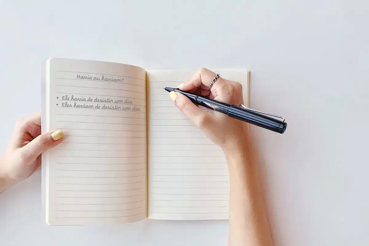 Escrevendo sobre a diferença entre Havia e Haviam. Pessoa escrevendo em um caderno com caneta preta, focando na diferença entre 'Havia' e 'Haviam'.