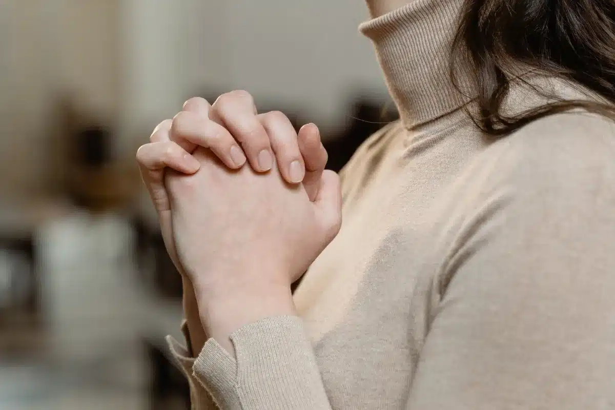 Mãos de uma pessoa entrelaçadas em oração, com fundo desfocado em ambiente interno.