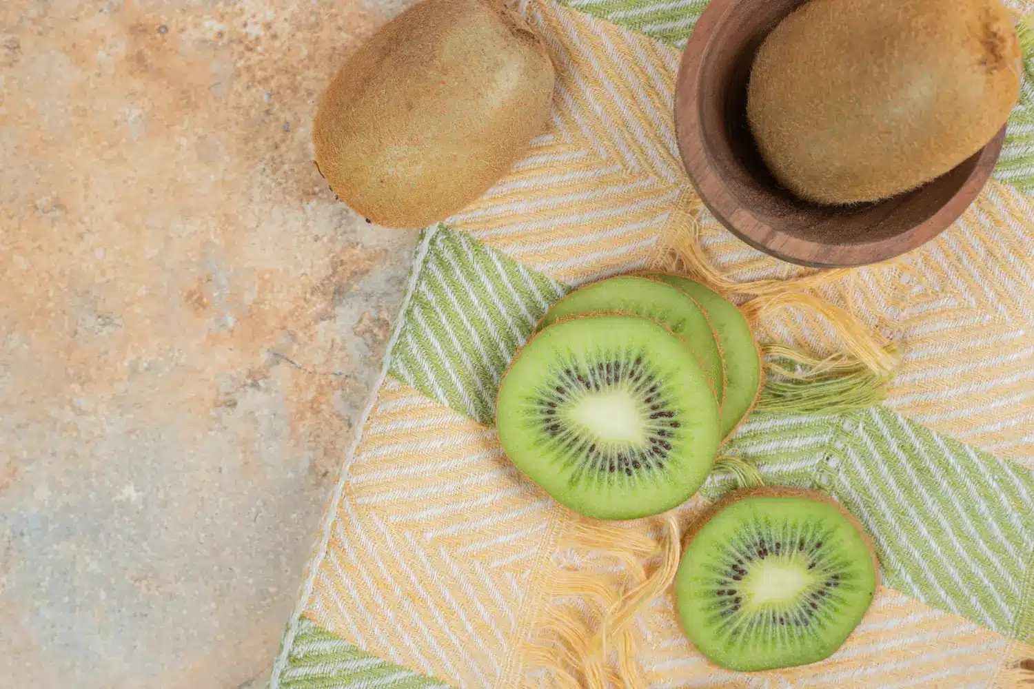 Kiwi cortado em fatias finas, disposto sobre um pano colorido com uma peça inteira ao lado.