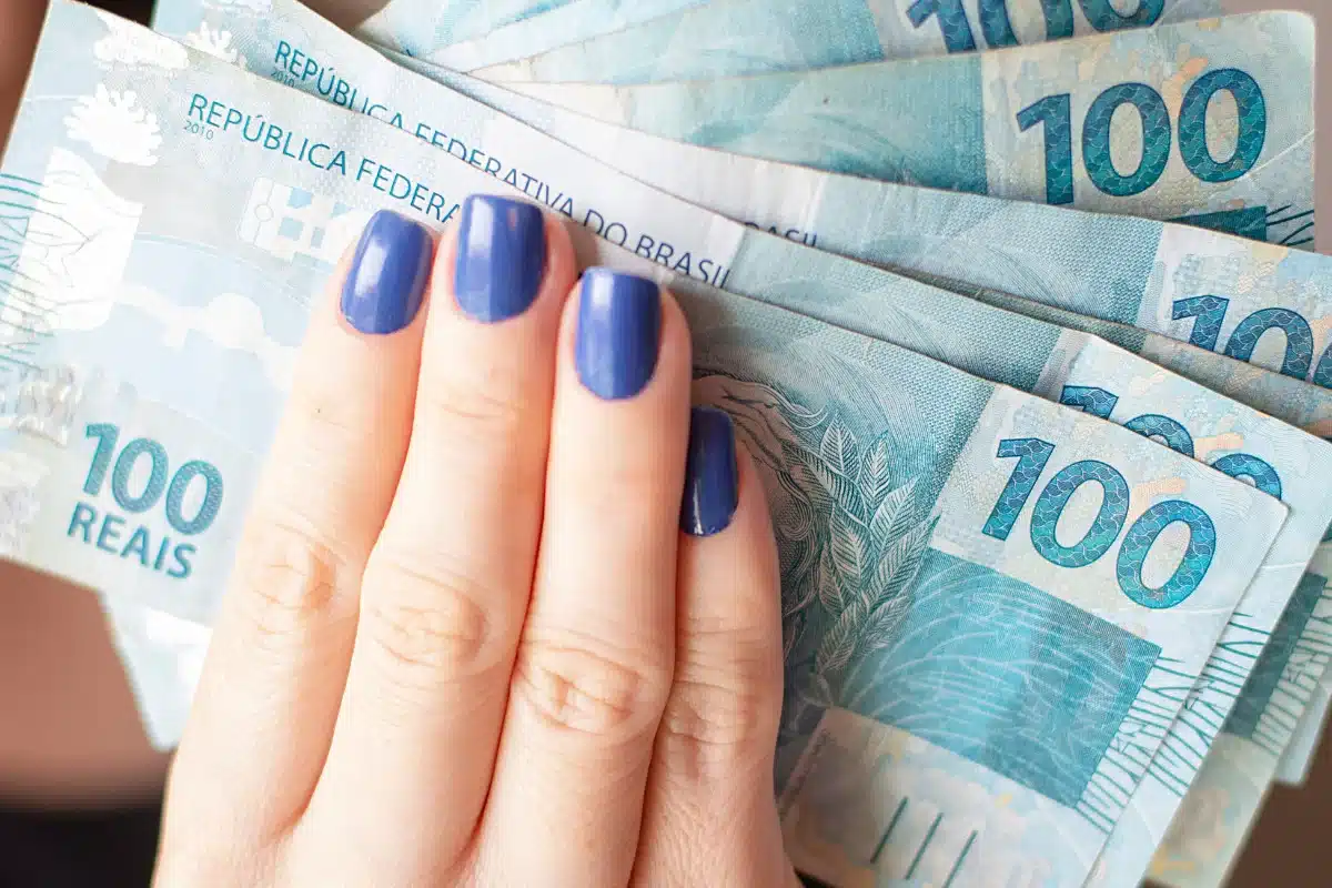 mão feminina com unhas pintadas segurando várias notas de 100 reais.