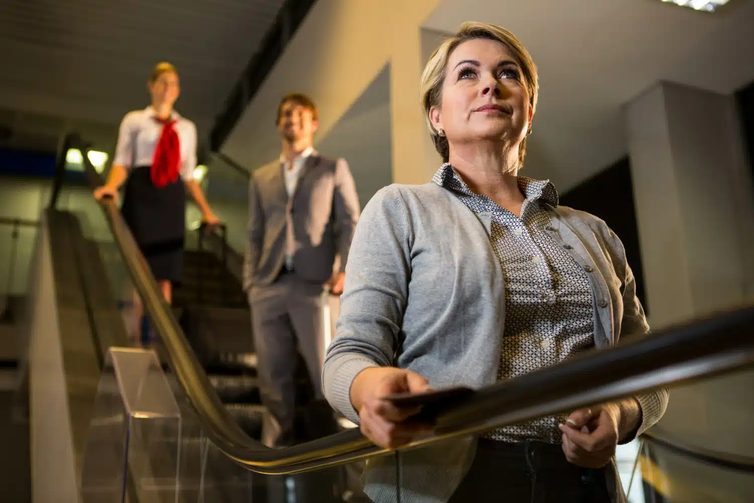 Mulher segura corrimão da escada rolante com dois profissionais ao fundo em ambiente corporativo.
