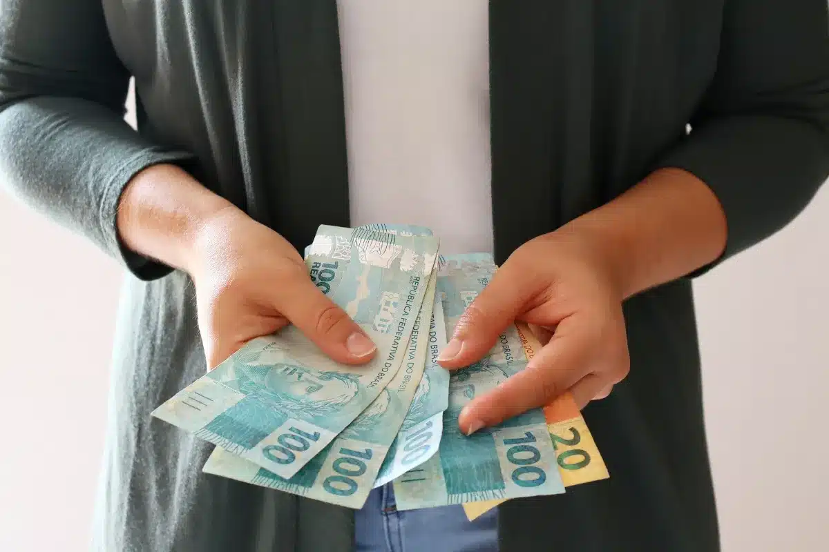 Pessoa segurando notas de R$100 e R$20, representando dinheiro em espécie.