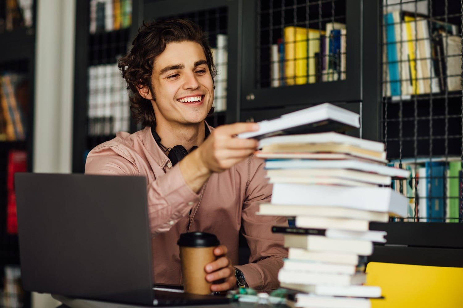 Jovem sorrindo em uma mesa cheia de livros, com um laptop aberto e um café em mãos, enquanto organiza o conteúdo de estudo para o Enem.