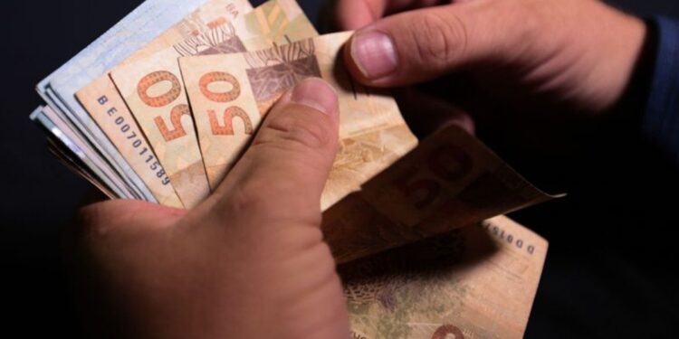 Imagem ilustrativa de mãos contando cédulas de cinquenta reais, em referência ao pagamento do INSS de julho