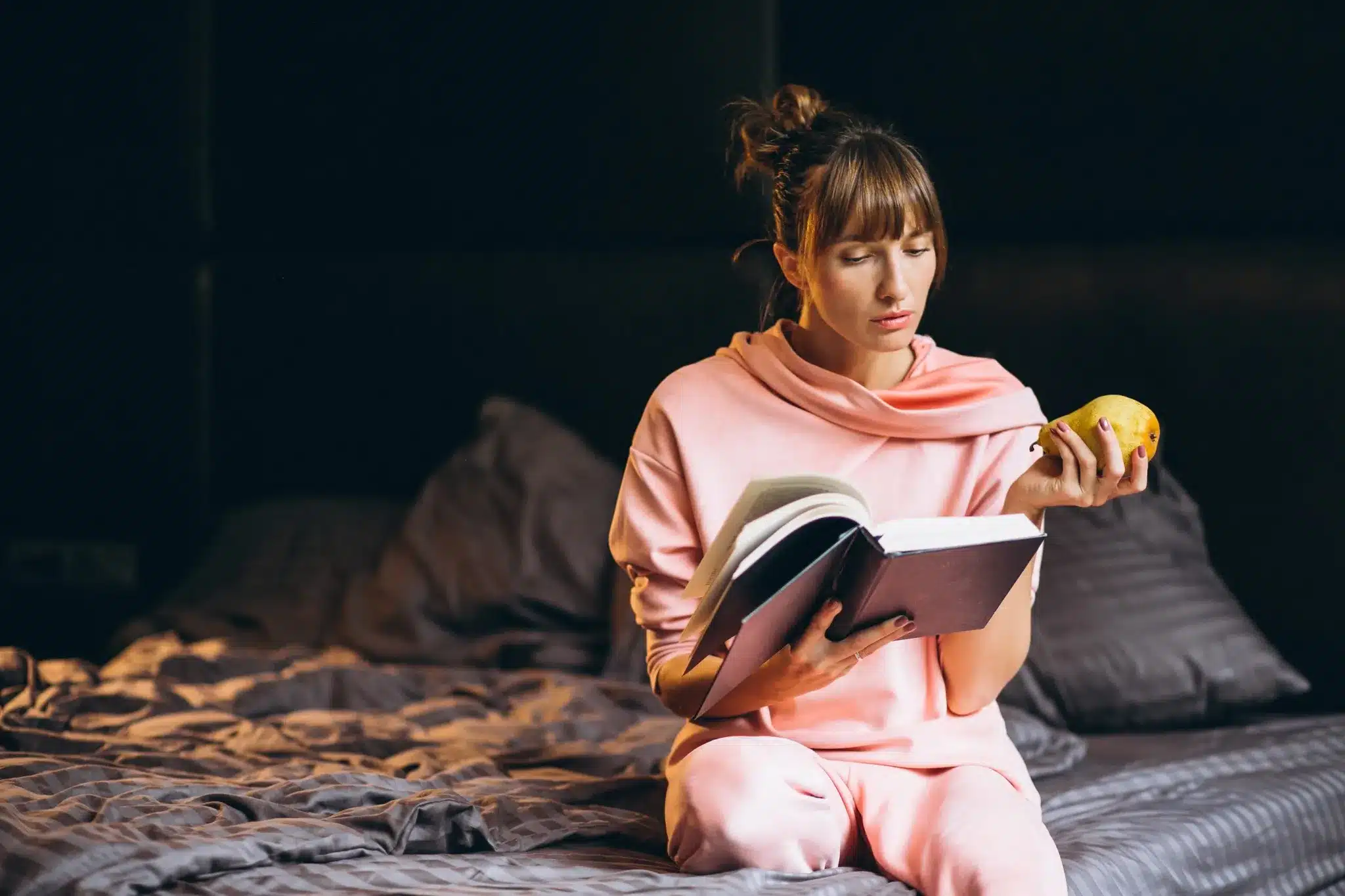 Mulher em pijama lendo livro enquanto segura uma pera na cama, sugerindo um momento de leitura em casa.