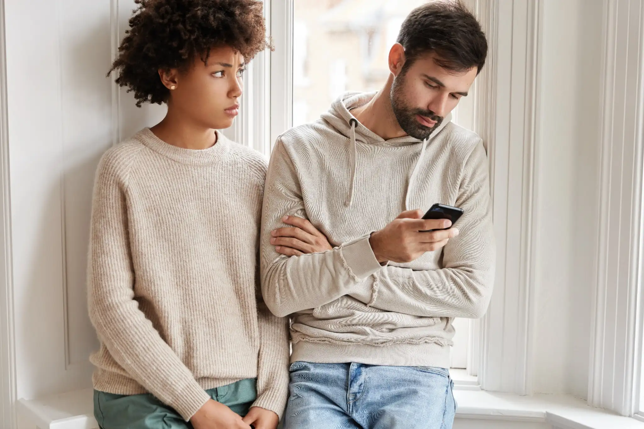 Sinais de indiferença Mulher triste em suéter observando seu namorado distraído no celular, refletindo sinais de indiferença no relacionamento.