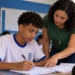 Professor orienta estudante em sala de aula, representando a regra de frequência mínima de 80% do Pé-de-Meia para receber o pagamento mensal.