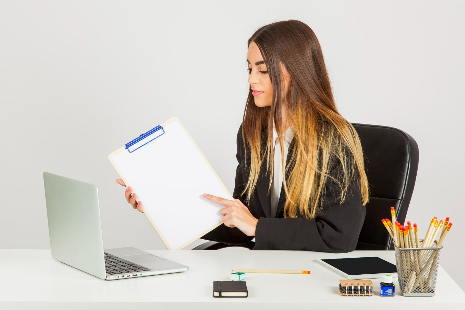 Mulher em traje formal mostrando uma prancheta vazia sentada à mesa de escritório com laptop e material de escritório.
