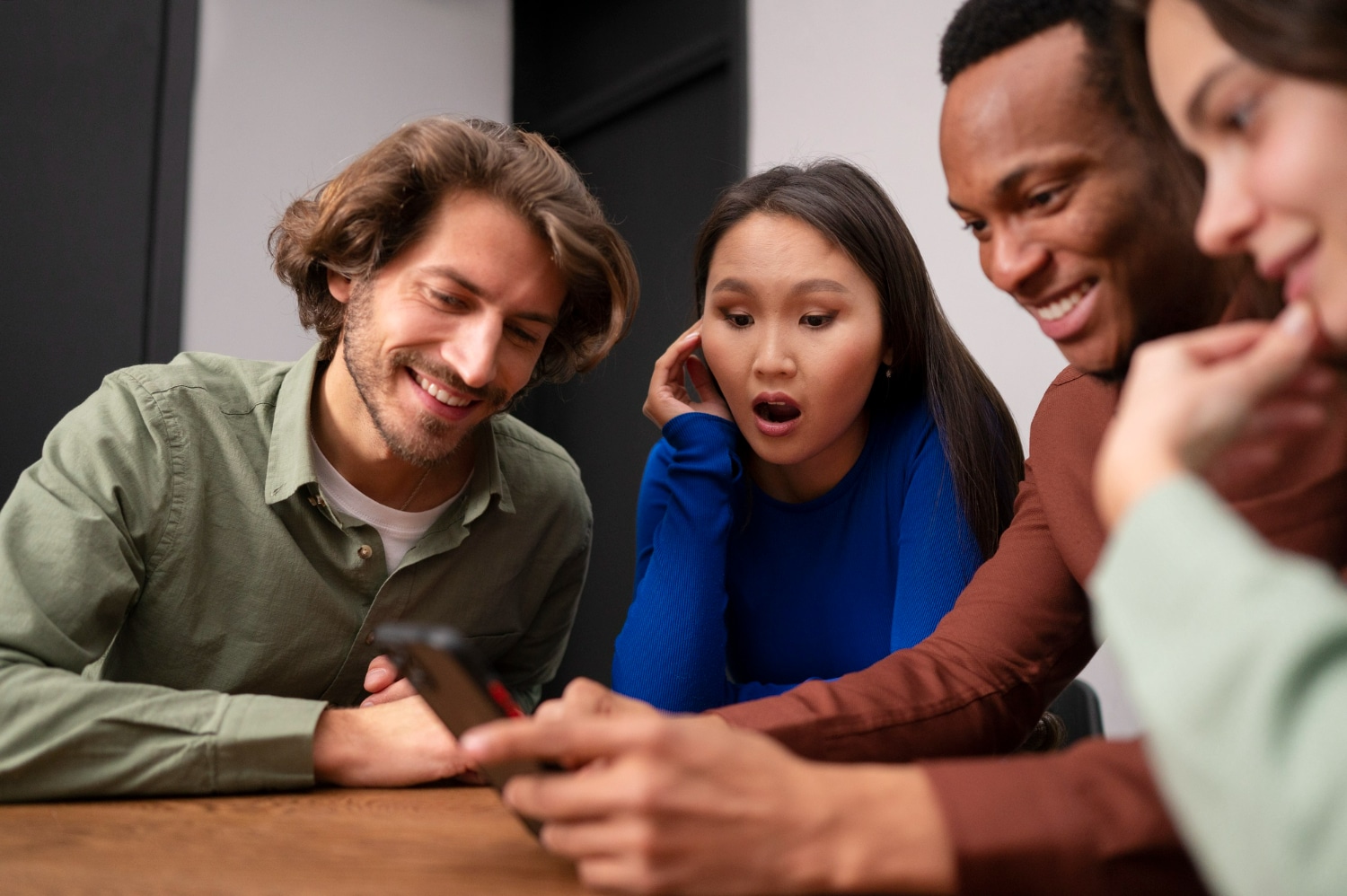 Grupo de quatro pessoas de diferentes etnias olhando para um celular com expressões variadas de surpresa e interesse.