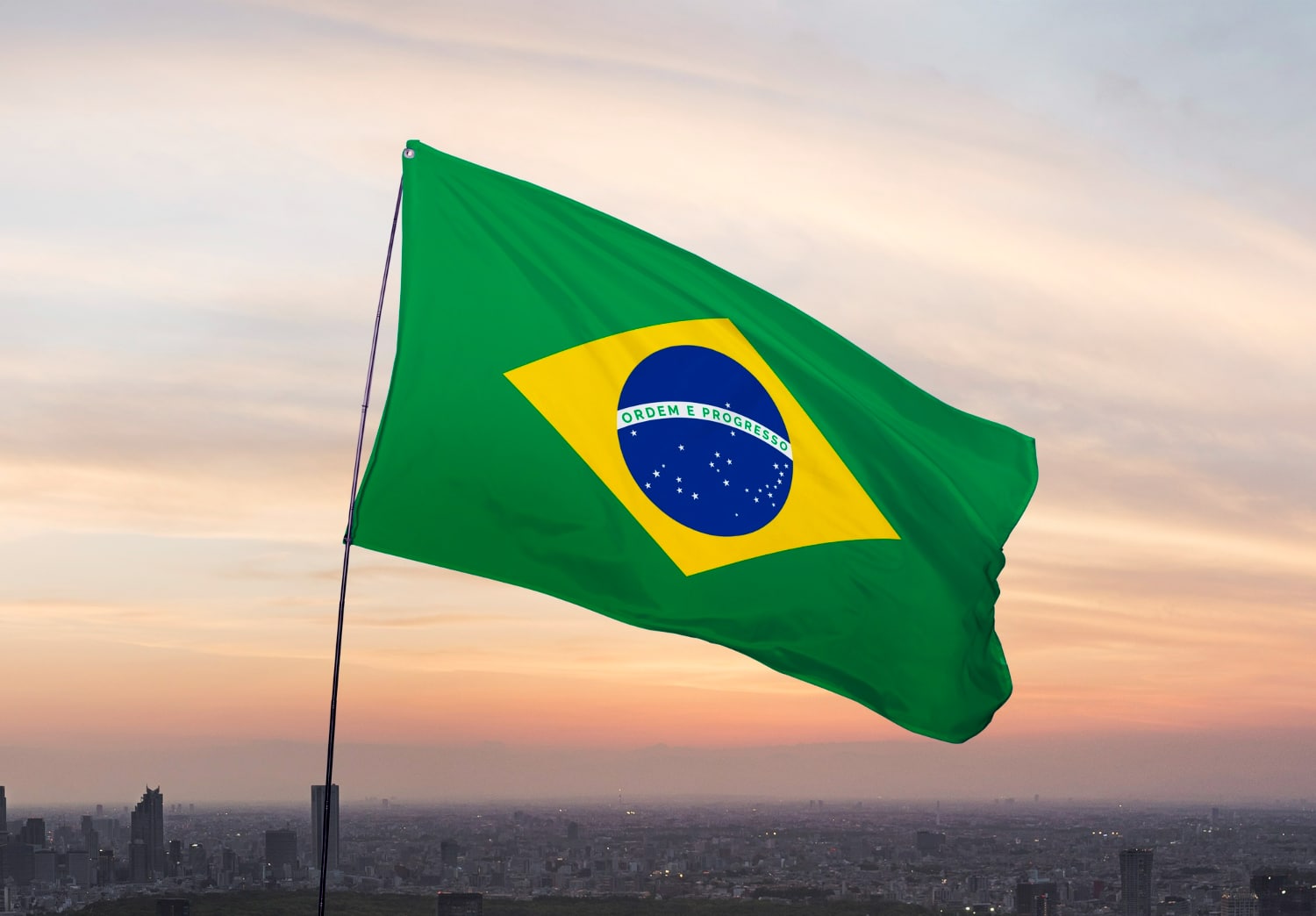 Bandeira do Brasil verde com losango amarelo e círculo azul com estrelas tremulando ao pôr do sol.