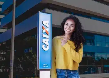 Mulher sorridente com blusa amarela e calça jeans, apontando para a placa da Caixa Econômica Federal, simbolizando o anúncio do novo concurso público e o andamento do processo seletivo