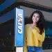Mulher sorridente com blusa amarela e calça jeans, apontando para a placa da Caixa Econômica Federal, simbolizando o anúncio do novo concurso público e o andamento do processo seletivo