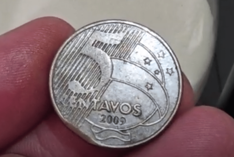 Moeda de 5 centavos de 2009: o raro defeito do disco cortado Uma mão segurando uma moeda de 5 centavos do ano de 2009 com um defeito na borda, o que a torna valiosa para colecionadores. A imagem serve para ilustrar um dos erros de cunhagem.
