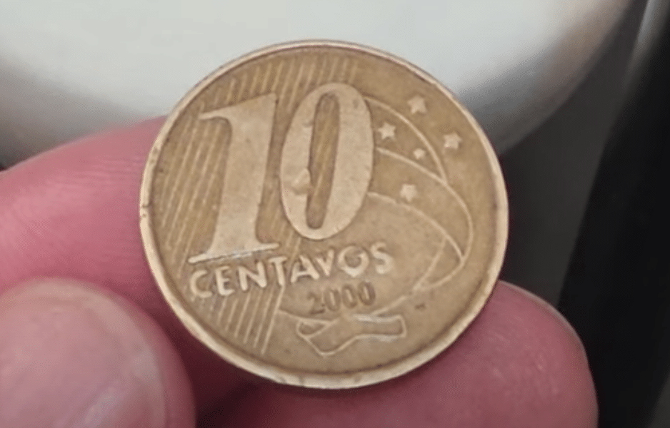 Moeda de 10 Centavos de 2000 com Erro de Cunhagem. Uma moeda de 10 centavos do ano 2000, segurada entre os dedos de uma pessoa, destacando a cunhagem com um possível defeito de fabricação.