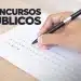 Pessoa preenchendo gabarito de prova de concurso público com caneta. O papel tem a inscrição 'CONCURSOS PÚBLICOS'.