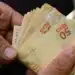 Mãos masculinas segurando várias notas de cinquenta reais em leque, fundo escuro, simbolizando os pagamentos do Pé-de-Meia de outubro.