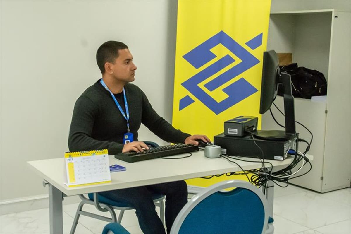 Salário inicial: Caixa ou Banco do Brasil?? Funcionário do Banco do Brasil trabalhando em computador na agência com uniforme e crachá da instituição