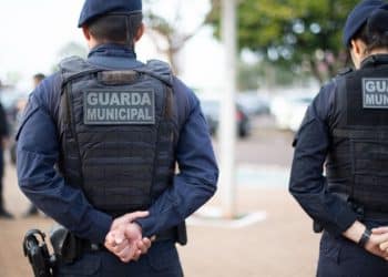 Dois guardas municipais com coletes pretos e uniformes azuis estão de costas em uma área externa urbana