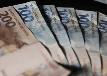Cédulas de real brasileiro em valores de 100 e 200 distribuídas sobre fundo escuro.