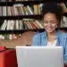 Mulher sorridente usando um laptop em um ambiente de biblioteca, representando o crescimento do número de alunos em EAD.