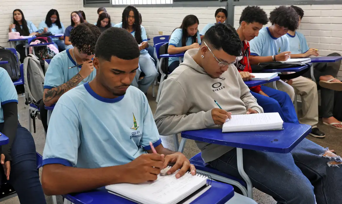 Pagamento pé-de-meia Jovens estudantes em sala de aula escrevendo em cadernos, com uniformes azuis e ambiente escolar organizado