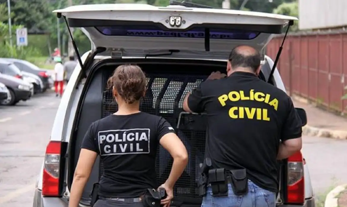 Delegado de Polícia Civil: Carreira, Salário e Atribuições Dois agentes da Polícia Civil, um homem e uma mulher, vistos de costas, em frente à parte traseira de uma viatura, participando de uma operação policial na rua.