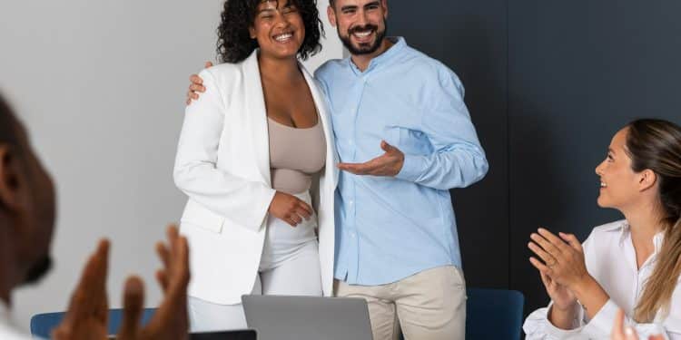 Duas pessoas sorridentes se abraçando e sendo aplaudidas por colegas em reunião profissional.