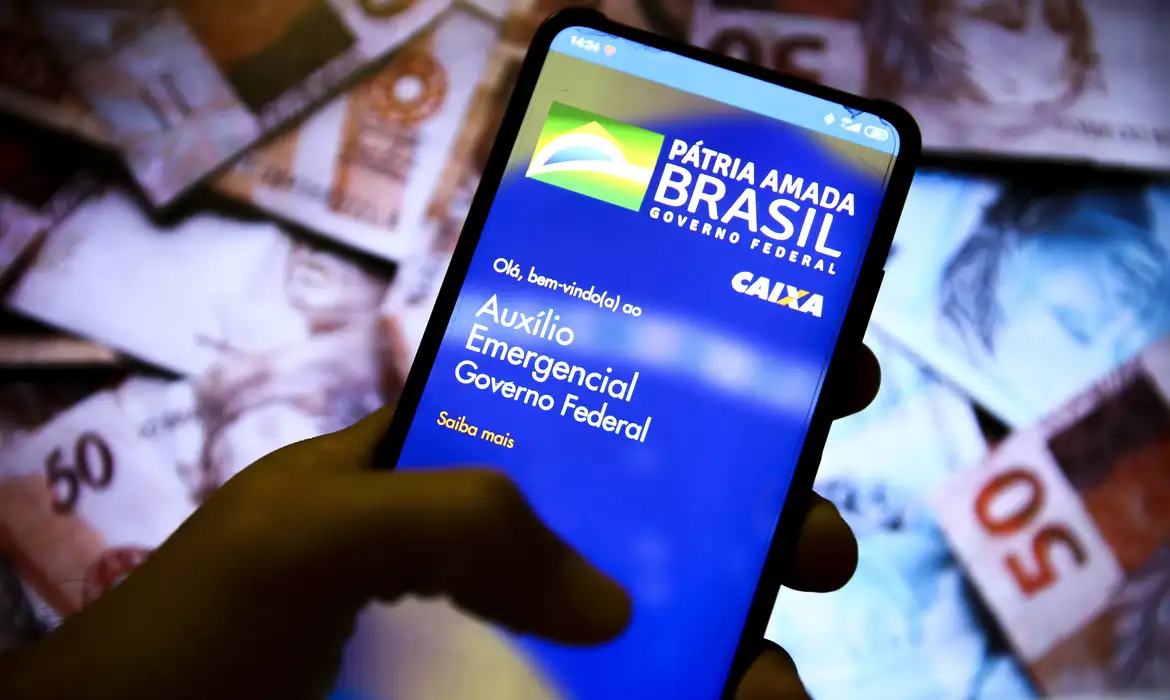 Devolução do Auxílio Emergencial: Quem é Isento e Não Precisa Devolver o Benefício Tela de celular exibindo o aplicativo do Auxílio Emergencial Caixa e notas de R$ 50 no fundo, ilustrando a distribuição do benefício e a polêmica sobre quem não precisa devolver.