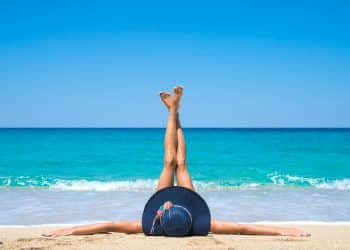 Pessoa deitada na areia da praia com chapéu azul e pernas esticadas para cima na frente do mar azul.
