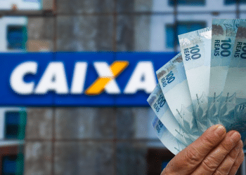 Mão segurando várias cédulas de 100 reais com o logo da Caixa Econômica ao fundo desfocado.