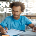 Jovem vestindo camiseta azul estudando para concursos em mesa com livros e caderno.