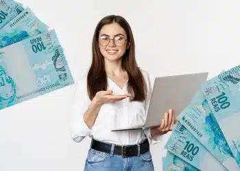 Mulher sorridente com óculos segurando laptop cercada por cédulas de 100 reais
