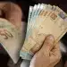 Mãos segurando várias cédulas de dinheiro em reais, incluindo notas de 5 e 50 reais, em foco próximo