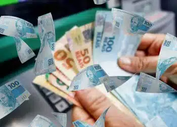 Mãos seguram diversas notas de 100 e 50 reais, enquanto outras cédulas de R$ 100 voam ao redor.