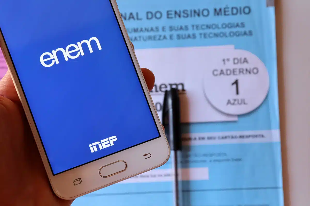 Enem Tela do aplicativo ENEM aberto em smartphone com caderno azul do exame e caneta ao fundo.