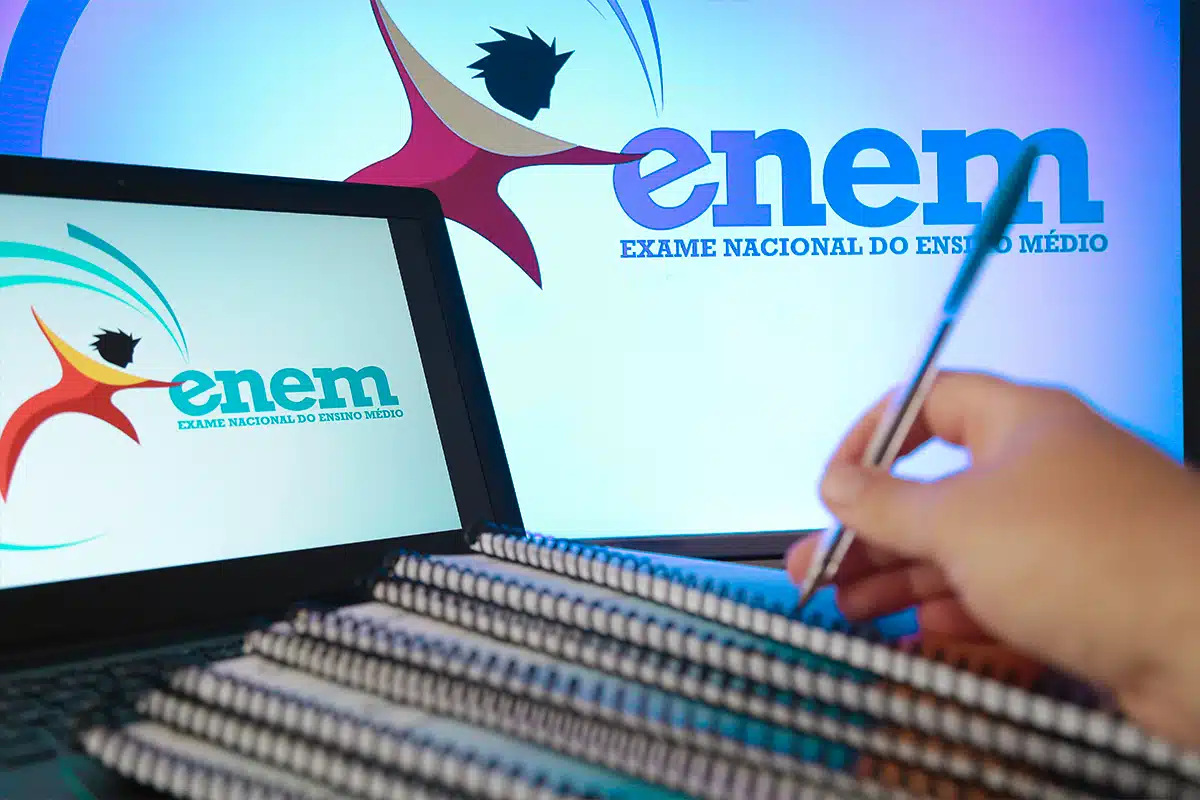 Estudante se preparando para o ENEM com anotações Mão escrevendo em caderno com logos do ENEM em tela de computador e monitor ao fundo