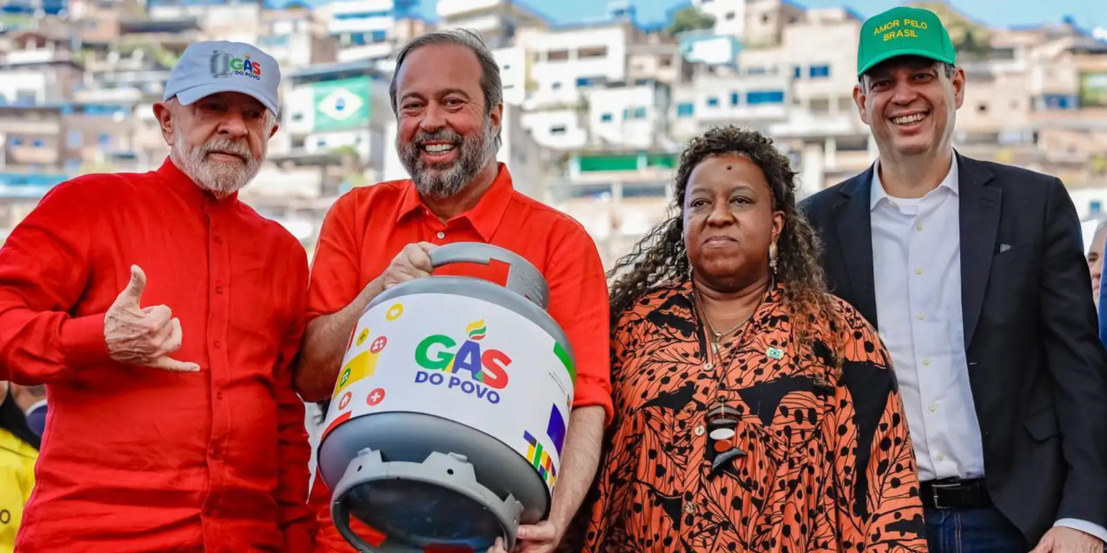Distribuição do projeto Gás do Povo com autoridades Quatro pessoas, duas com camisas vermelhas, segurando botijão de gás do projeto Gás do Povo em comunidade