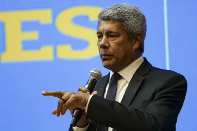 Homem falando em microfone durante palestra formal Homem de terno preto segurando microfone e gesticulando durante discurso em fundo azul
