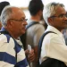 Grupo de pessoas em fila, incluindo homens com cabelo grisalho e óculos, esperando em ambiente fechado