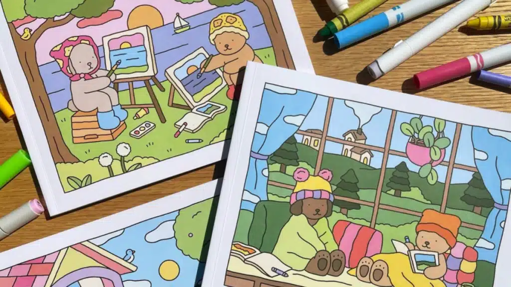 Livros de Colorir Bobbie Goods: A Arte Terapêutica de Abbie Gouveia Livros de colorir abertos com ilustrações adoráveis de ursinhos e cachorrinhos em cenas tranquilas, rodeados por canetas e lápis de cor, destacando a arte da Bobbie Goods.