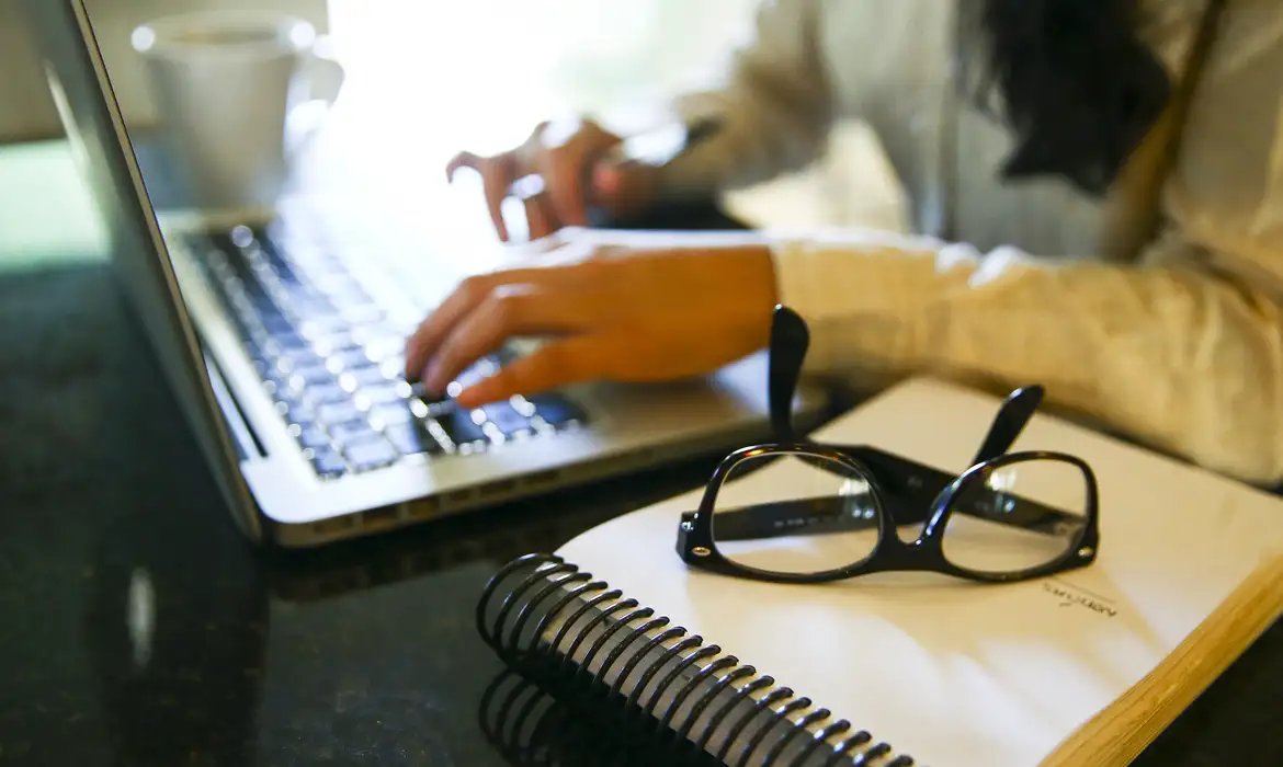 Pessoa digitando em laptop com caderno e óculos Mãos digitando em laptop ao lado de caderno espiralado branco e óculos de grau pretos sobre mesa preta